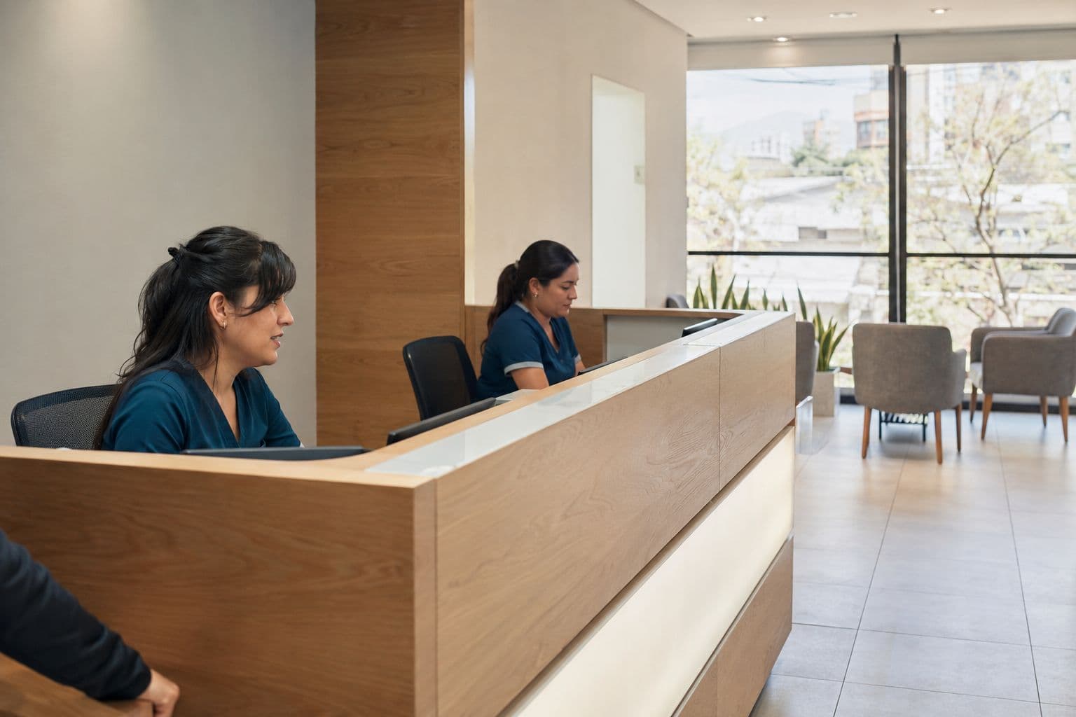 Recepción de una clínica con mostrador de madera clara, dos personas del equipo administrativo atendiendo y una sala de espera con ventanal al fondo
