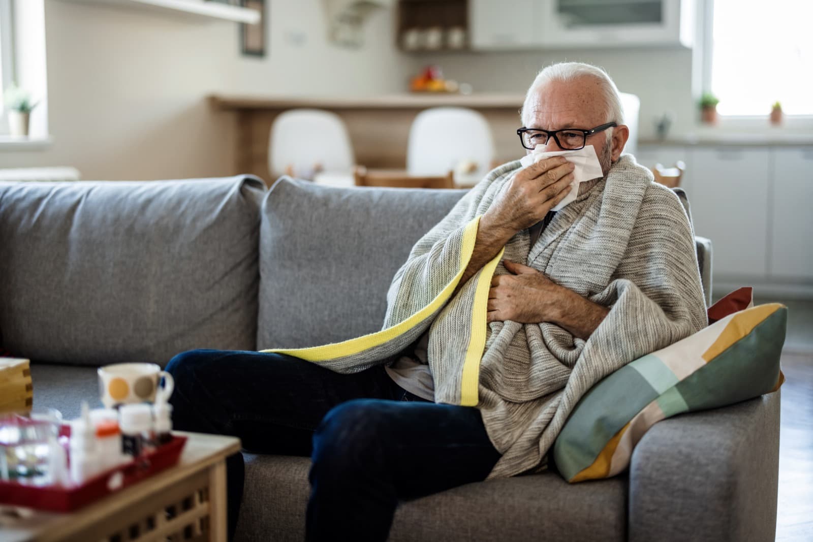 Hombre mayor con gripe, envuelto en una manta en el sillón de su casa, sonándose la nariz con un pañuelo, con medicamentos sobre la mesa
